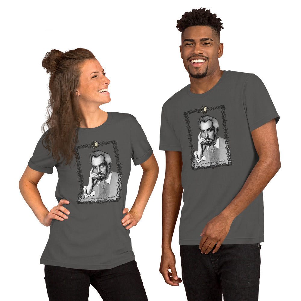 Two models wearing matching asphalt gray unisex t-shirts featuring a black-and-white vintage portrait illustration of a distinguished older man in an ornate decorative frame with a small cameo medallion at the top and text reading "Have you anything to say, warlock?" at the bottom. The woman on the left is laughing with her hair in a half-up style; the man on the right is smiling broadly. White background.