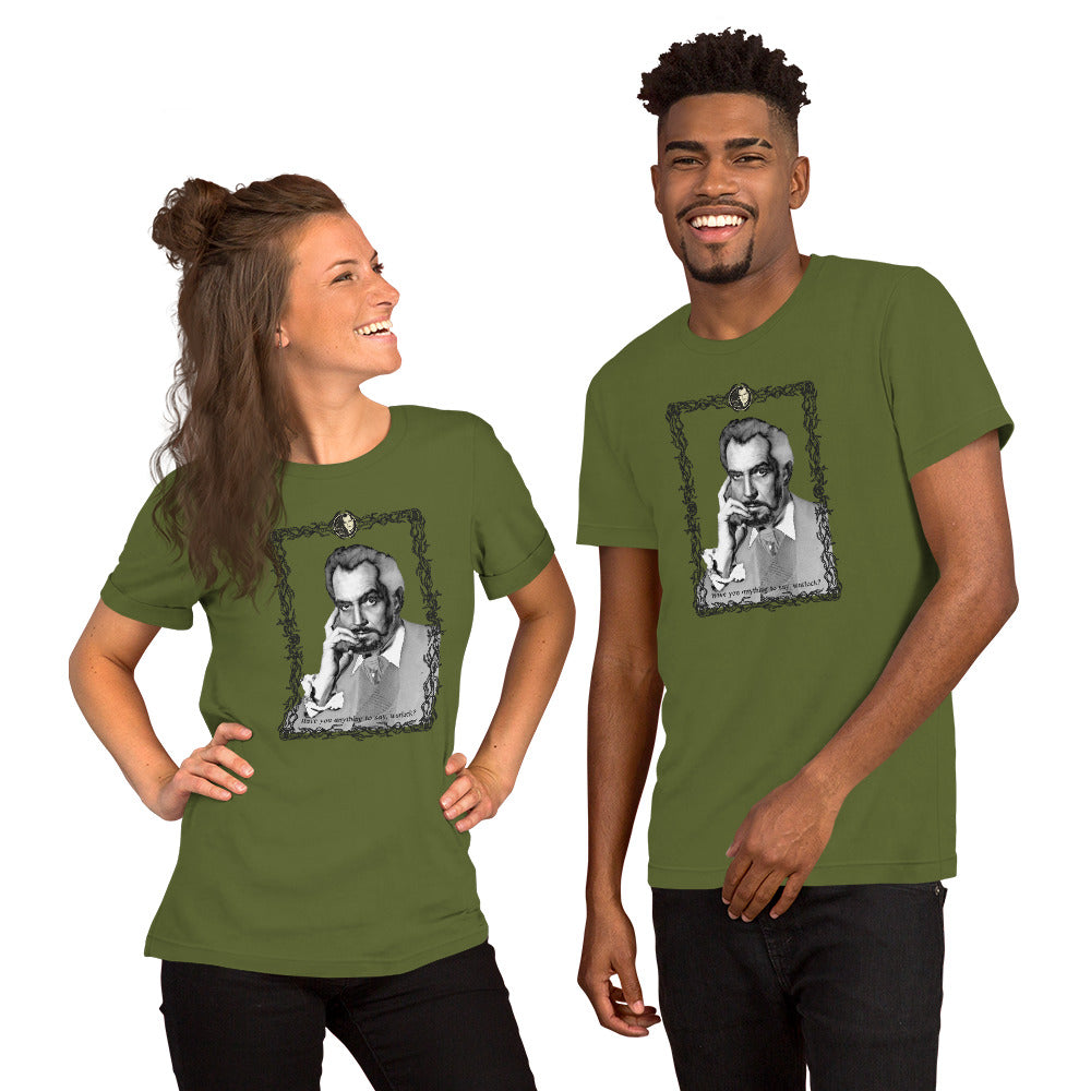 Two models wearing matching olive green unisex t-shirts featuring a black-and-white vintage portrait illustration of a distinguished older man in an ornate decorative frame with a small cameo medallion at the top and text reading "Have you anything to say, warlock?" at the bottom. The woman on the left is laughing with her hair in a half-up style; the man on the right is smiling broadly. White background.