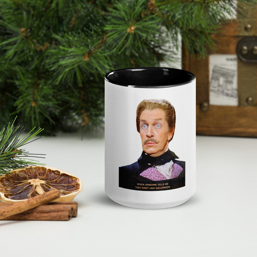 A ceramic mug with a black rim and handle, featuring a black and white photo of a person with exaggerated facial features labeled 'Vincent Price - The King of Horror', placed on a table with pine branches and cinnamon sticks in the background.